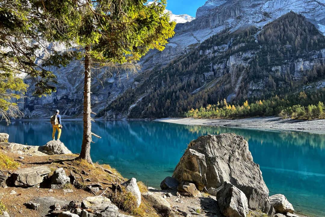 Excursionista sobre una roca a la orilla del Oeschinensee mirando el lago glaciar turquesa hacia los acantilados calizos del Blüemlisalp en otoño, Kandersteg, Suiza