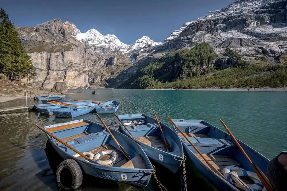 Barcas de remos azules de alquiler amarradas en el embarcadero del Oeschinensee con las cumbres nevadas del Blüemlisalp reflejadas en el agua turquesa, Kandersteg, Suiza
