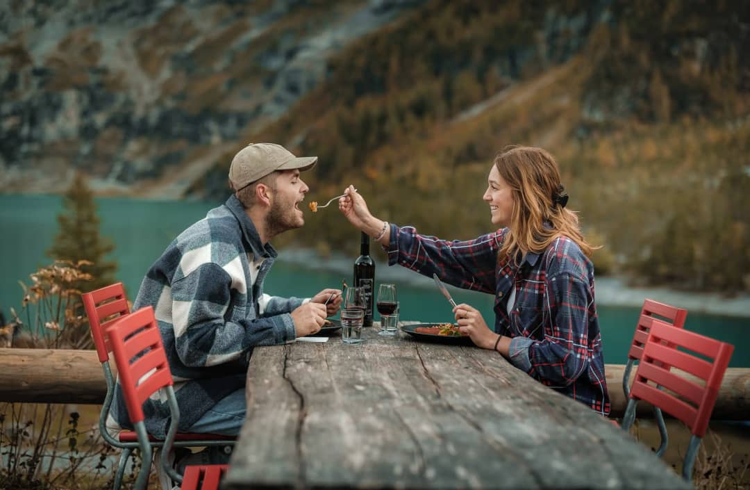 Pareja cenando en la terraza del Restaurante Oeschinensee con vino tinto y vistas al lago turquesa y al paisaje alpino de otoño, Kandersteg, Suiza