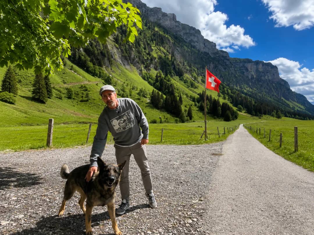 Guest walking with farm dog on the valley path to Alp Signswiler, Swiss flag and alpine cliffs in the background, Justistal farm tour from Interlaken