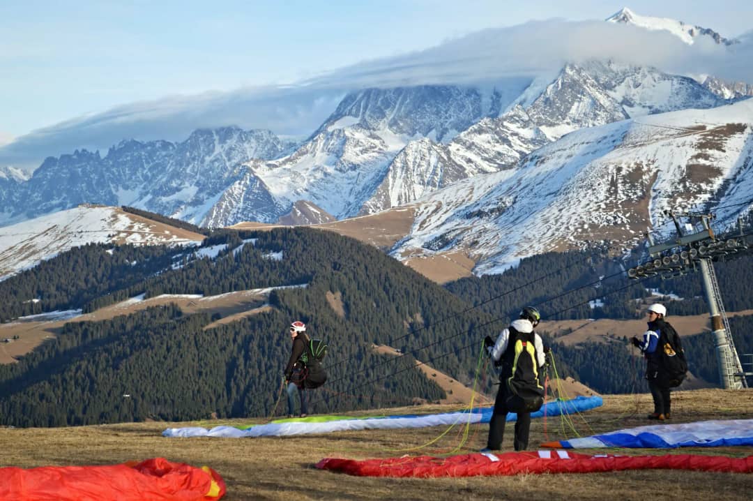 Paragliding pilots preparing canopies on alpine launch site with snow-capped Swiss Alps peaks and cable car in background