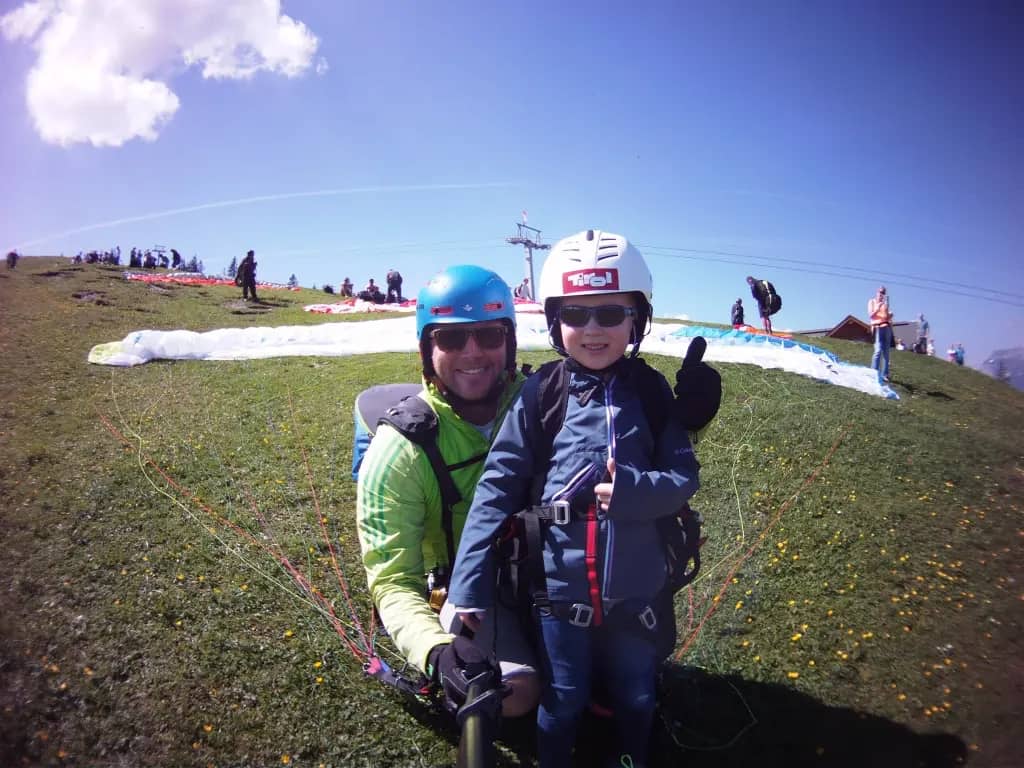 Young child with thumbs up in paragliding harness with pilot at alpine launch site, children tandem paragliding Switzerland