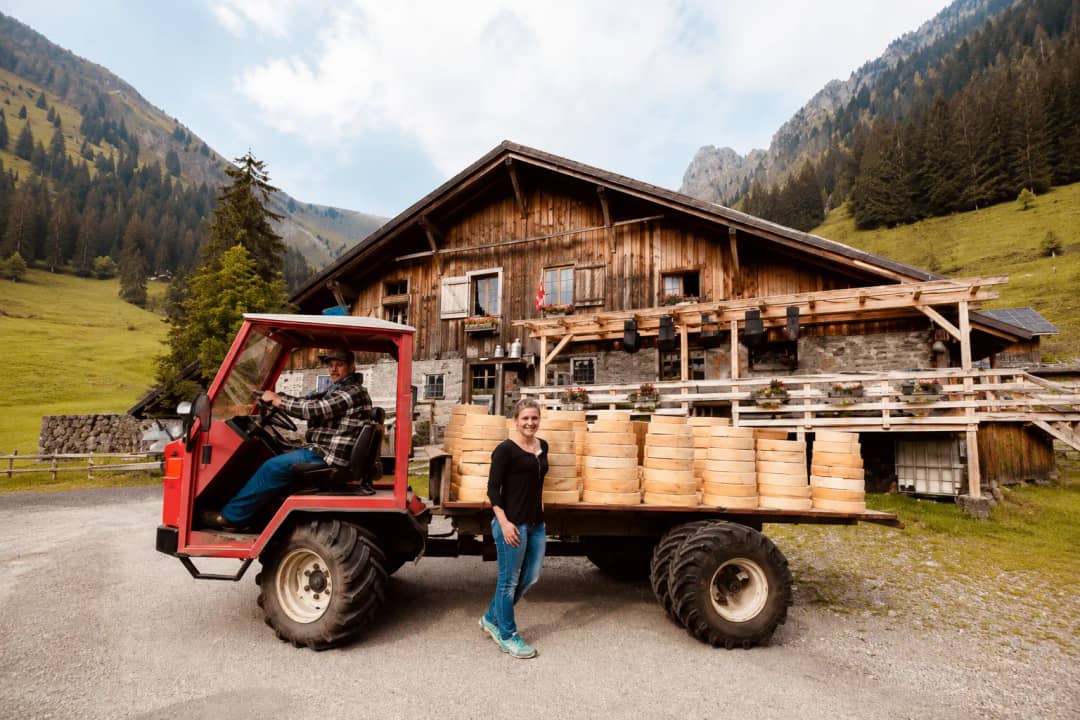 Daniela standing next to a farm vehicle loaded with stacks of artisanal Swiss cheese wheels, in front of a traditional wooden alpine chalet.