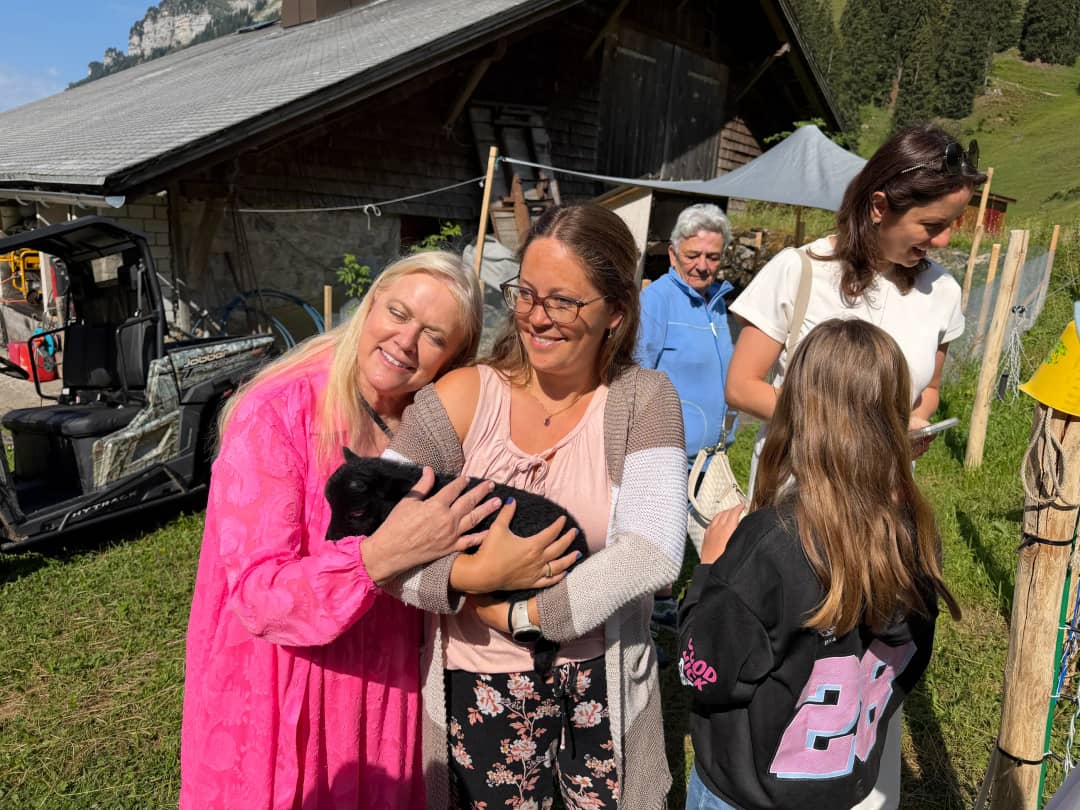 Family cradling a black baby goat outside the farmhouse while a girl reaches out to stroke it