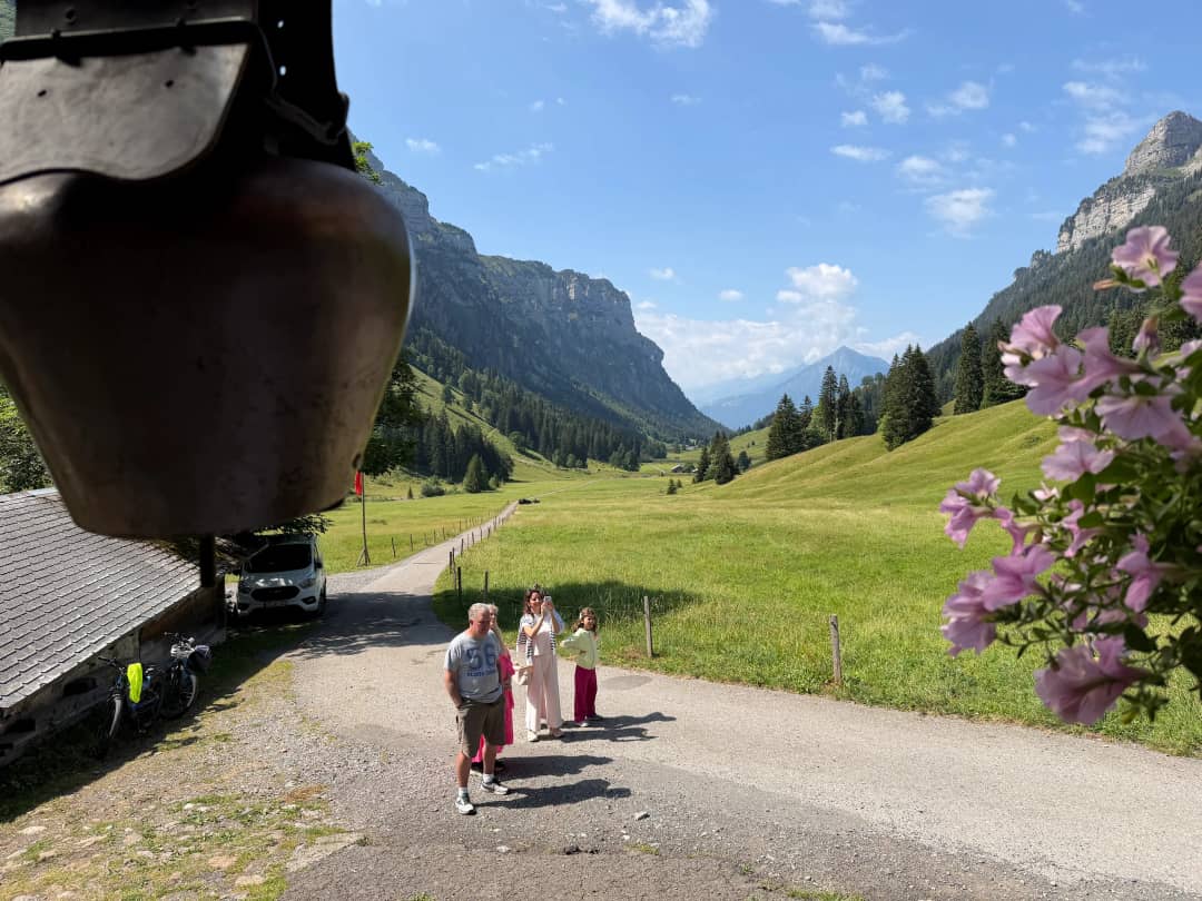 Grandparents, mother and daughter taking photos on the flat farm track in the Justistal, framed by a cowbell and pink flowers