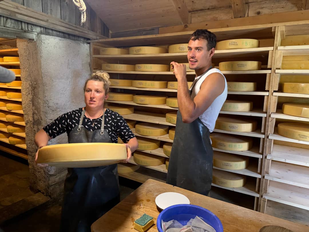 Daniela holding up a wheel of alpine cheese in the ageing cellar, shelves of Alpkäse stacked behind her