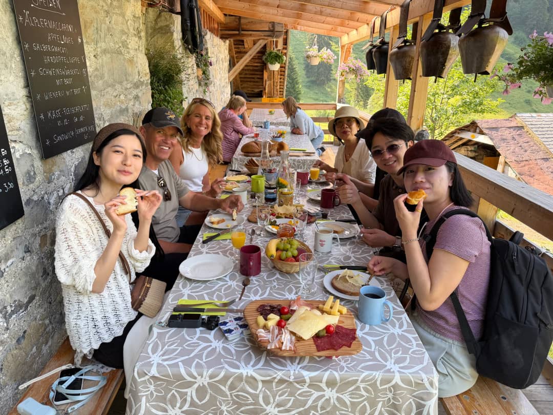 Guests sharing an alpine farm breakfast on a Swiss farm terrace, cheese, cured meat and bread from the same valley