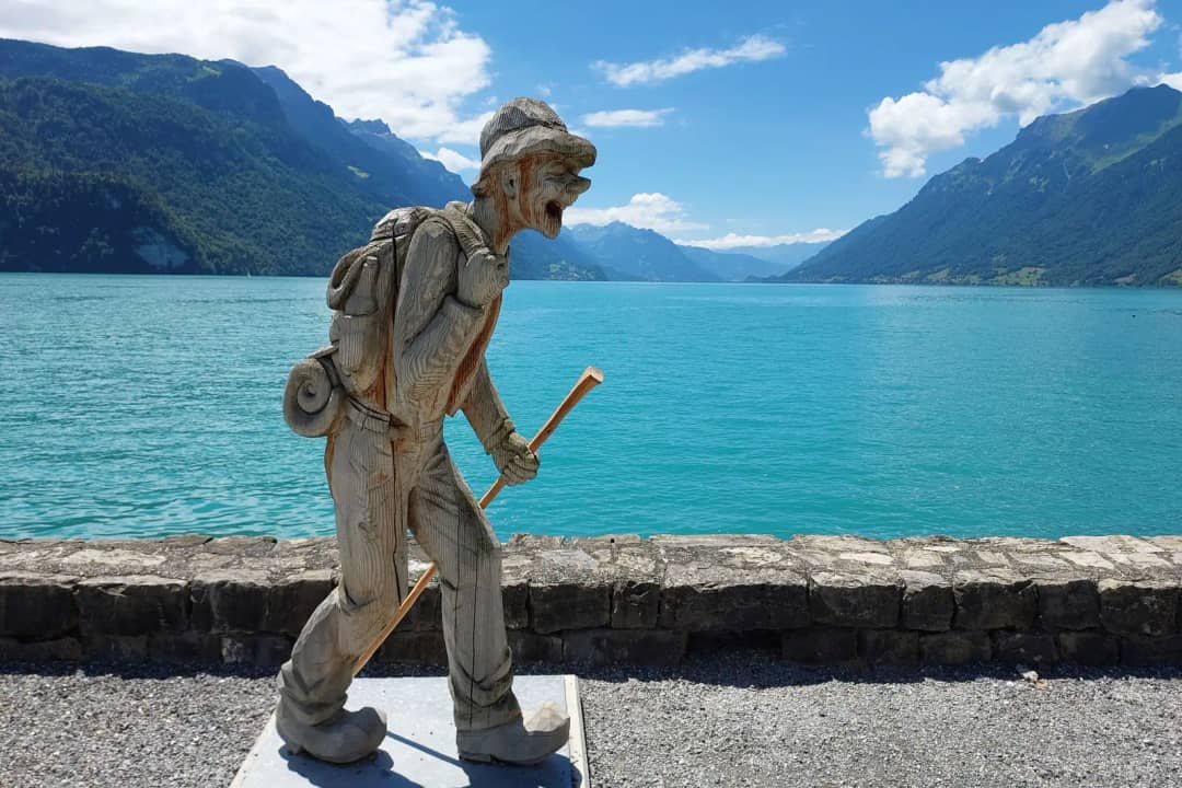 Geschnitzte Holzfigur eines Wanderers mit Stock auf einer Steinmauer am tĂĽrkisfarbenen Wasser eines Sees