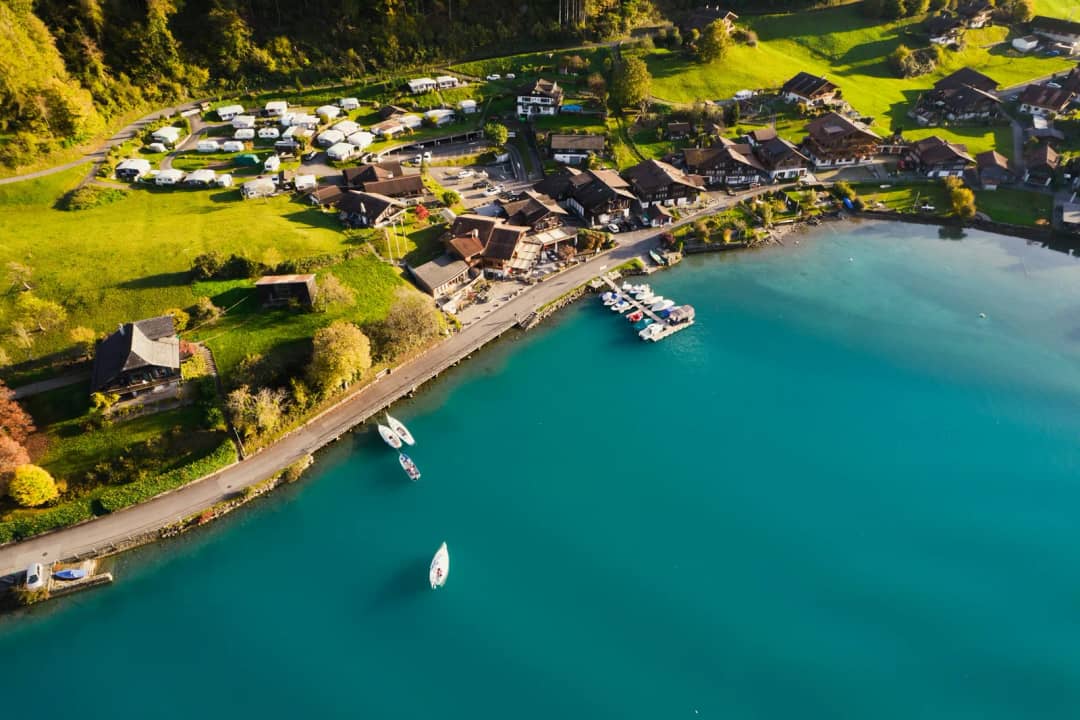 Luftaufnahme eines Dorfes mit Campingplatz und Schiffsanlegestelle am türkisfarbenen Ufer des Brienzersees