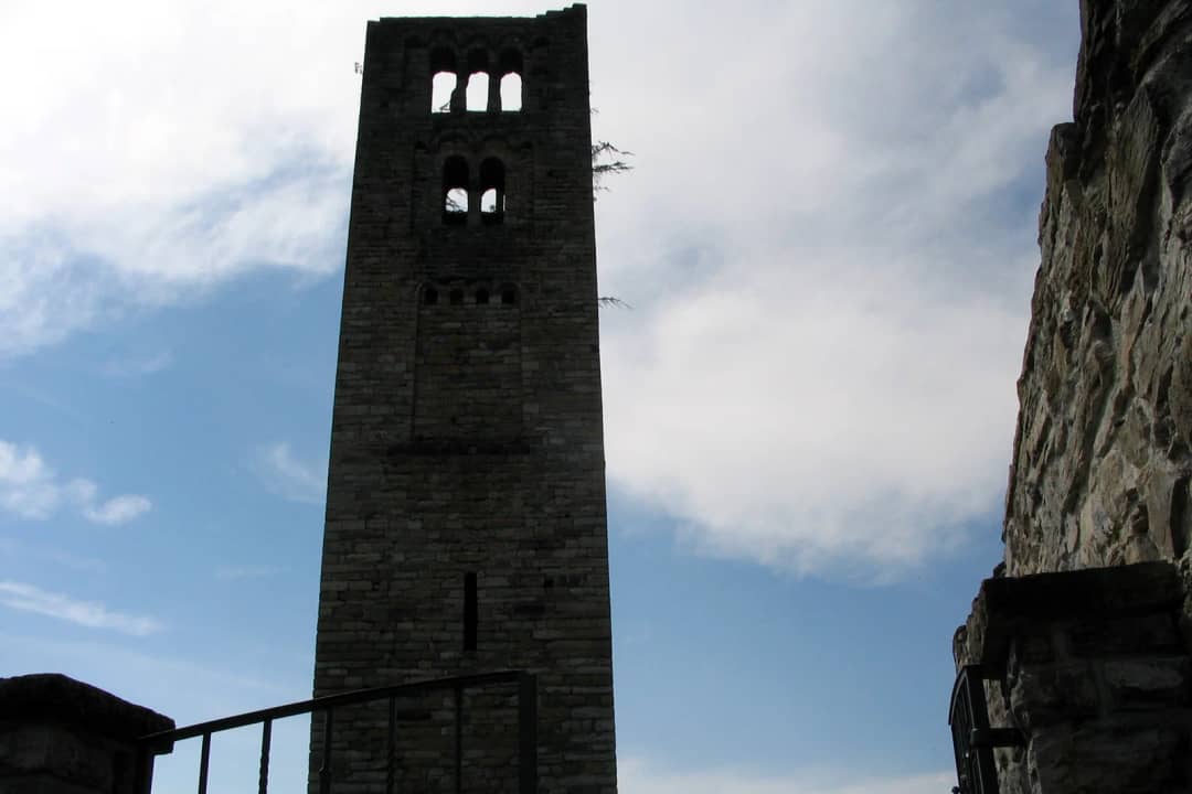 Stone bell tower of the Goldswil church ruin rising against a blue sky with clouds beside an old iron railing