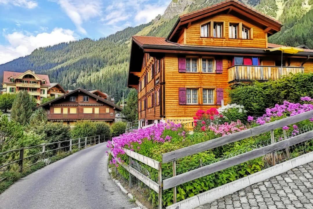 Gran chalet de madera con balcones sobre un parterre de flores y una valla de madera junto a un camino alpino