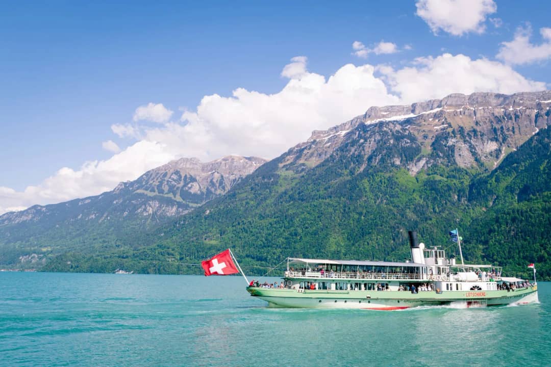 Raddampfer mit Schweizer Flagge auf einem türkisfarbenen See unter bewaldeten Hängen und Felsspitzen