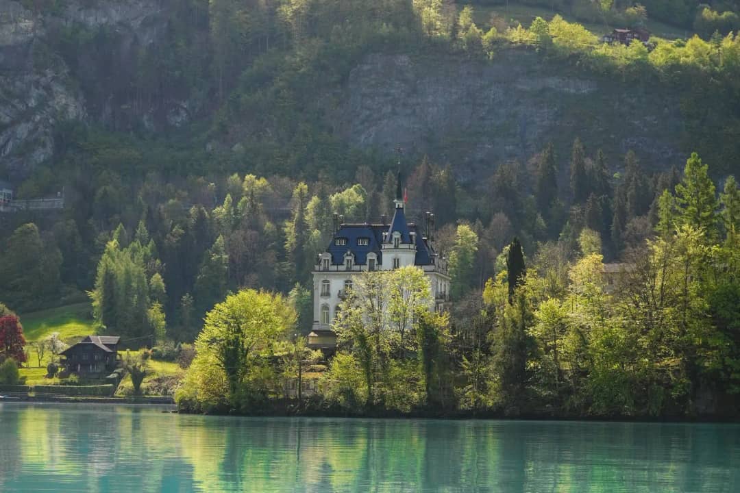 Manoir blanc aux airs de château parmi les arbres de la rive, reflété dans l'eau turquoise et calme du lac