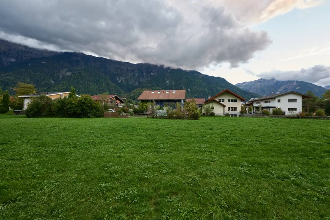 Weite grüne Wiese vor den Häusern von Bönigen unter bewölktem Himmel, dahinter bewaldete Berge