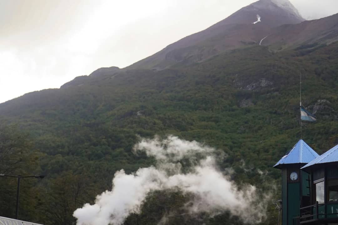 Weisser Dampf steigt neben dem Dach einer kleinen Bergstation auf, darüber ein grüner Berg in treibenden Wolken