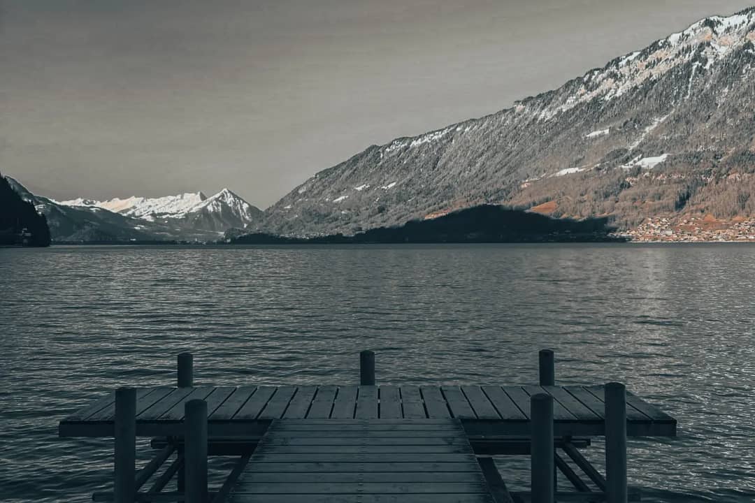 Ponton en bois avançant dans l'eau gris sombre d'un lac suisse sous des montagnes enneigées, ciel couvert