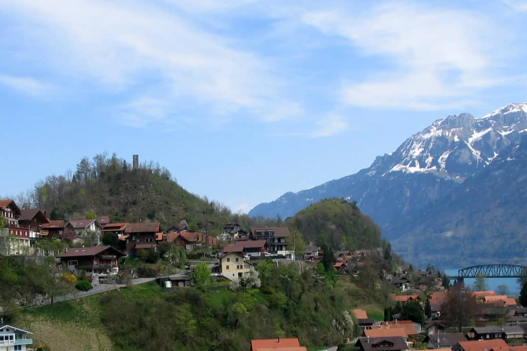 Houses of Goldswil on a green slope with a wooded hill and its ruined tower, snowy peaks rising behind