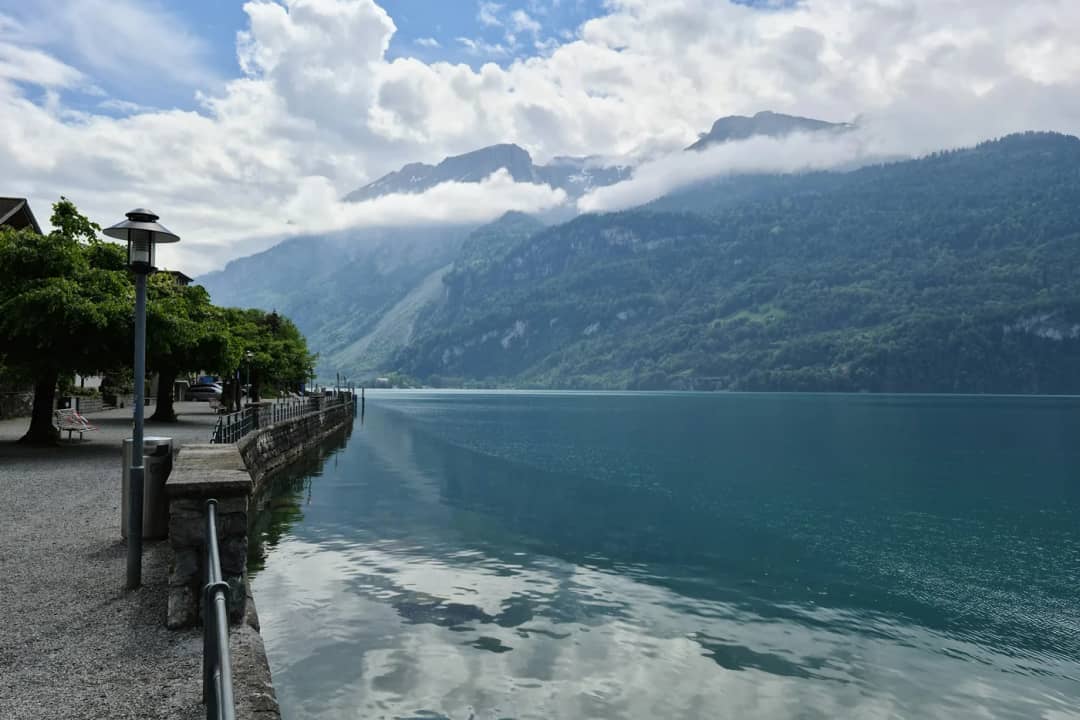Paseo junto al lago con farolas y agua tranquila que refleja nubes bajas y montañas en un día gris