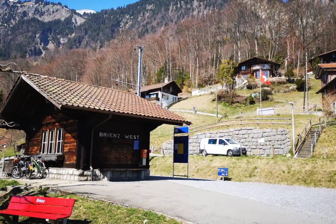 Pequeño edificio de madera de la estación de Brienz West junto a la carretera, con chalets en la ladera