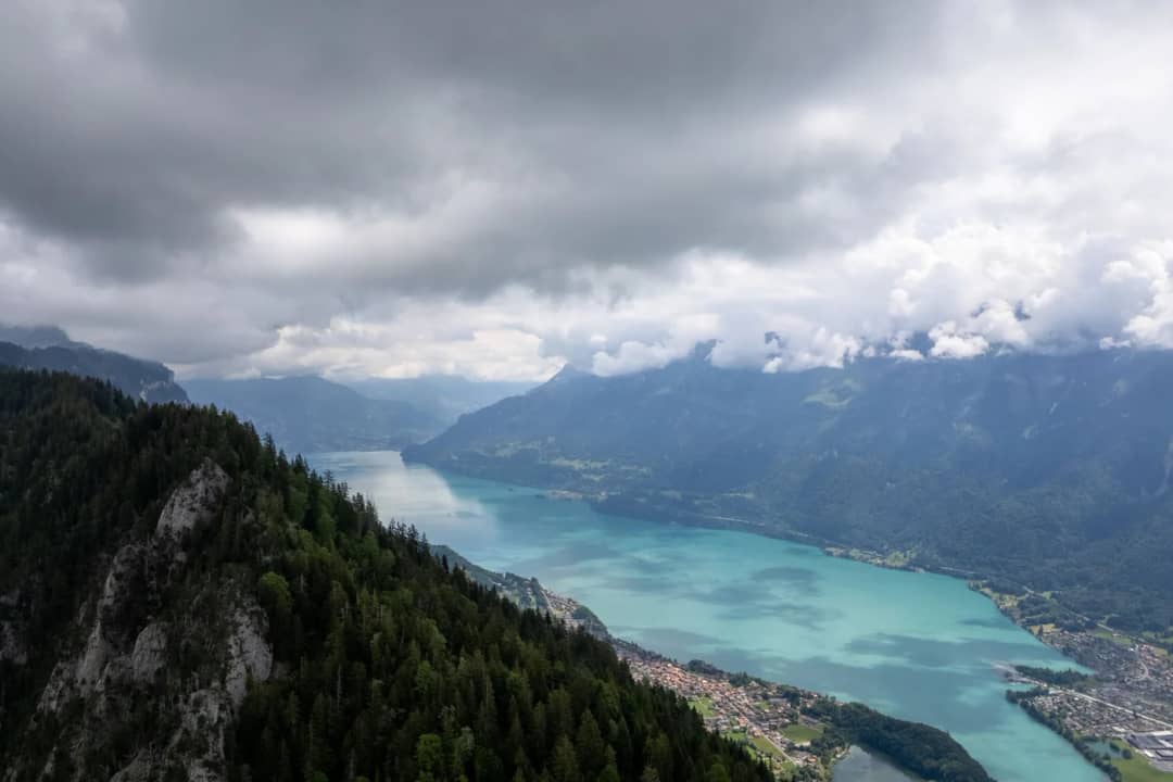 Türkisfarbener Bergsee zwischen dunklen, bewaldeten Hängen unter schweren grauen Wolken, von oben gesehen