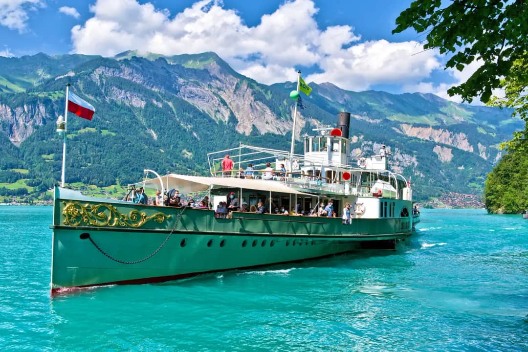 Raddampfer mit grünem Rumpf, Flaggen und Fahrgästen an Deck auf türkisfarbenem Wasser unter hohen Bergen