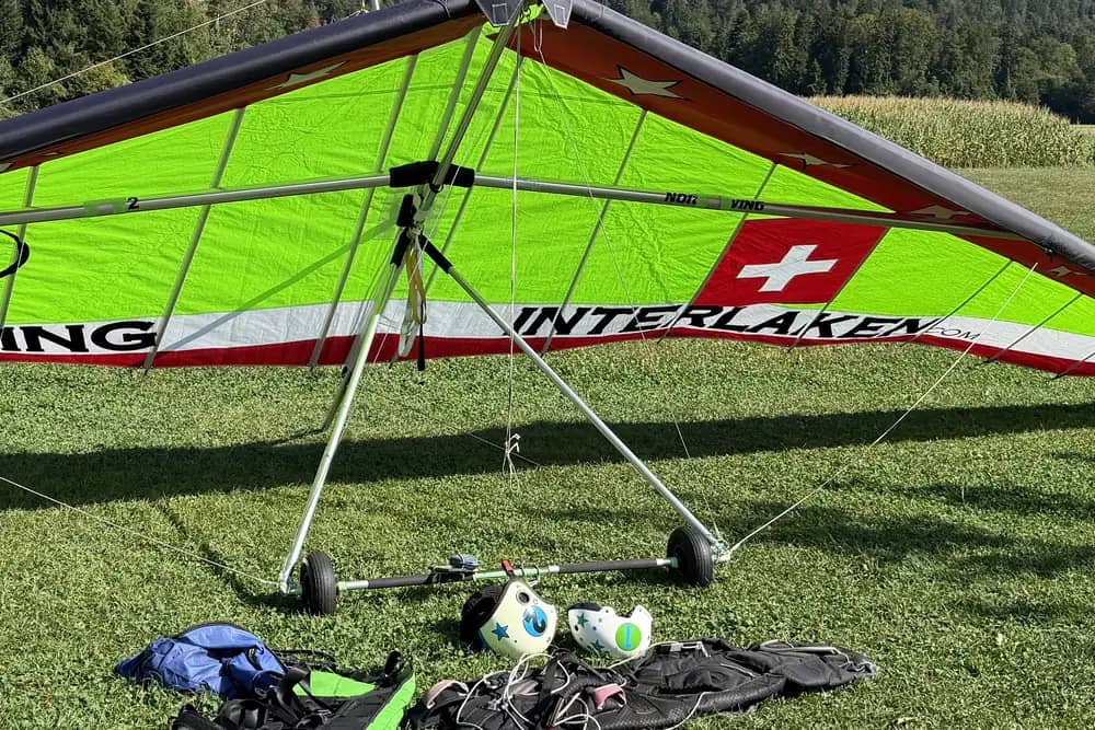 Starrer Steuerbügel mit Landerädern unter einem Tandem-Hängegleiter, daneben Gurtzeuge und Helme im Gras