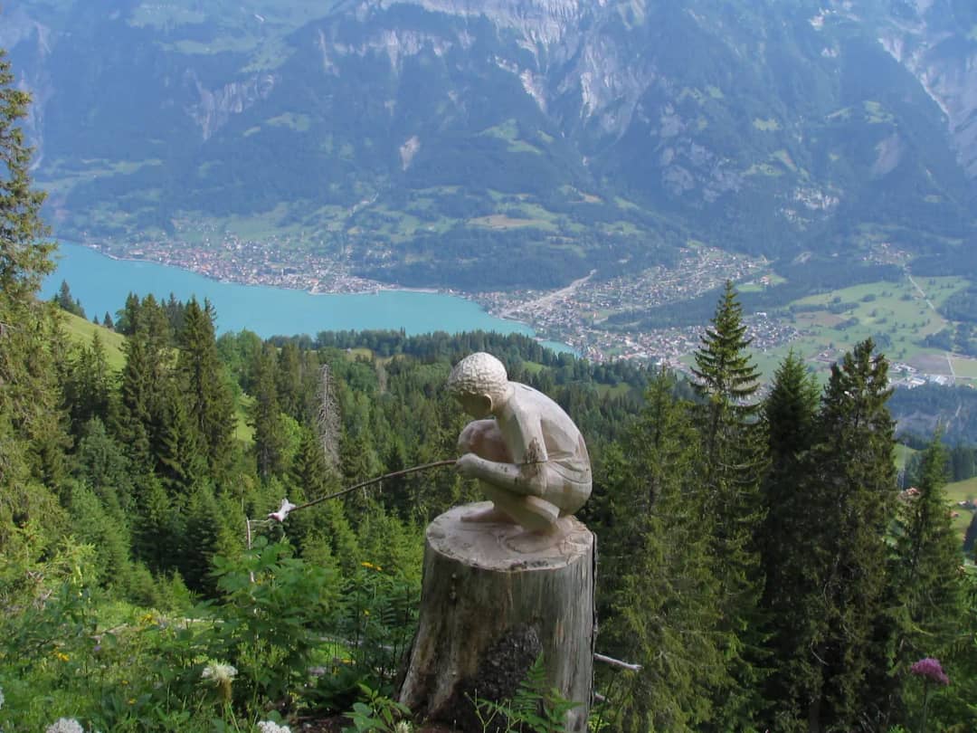 Holzskulptur eines Knaben, der eine Wurst brät, am Schnitzlerweg auf der Axalp, tief unten Brienz und sein See
