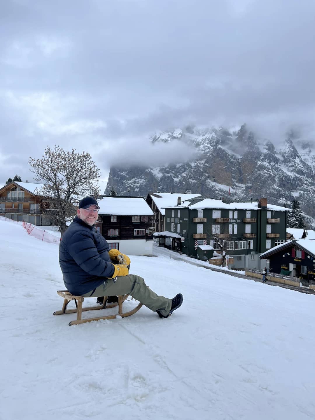 Mürren village in early April 2026, a sledder in the snow in front of chalets and hotels under low cloud