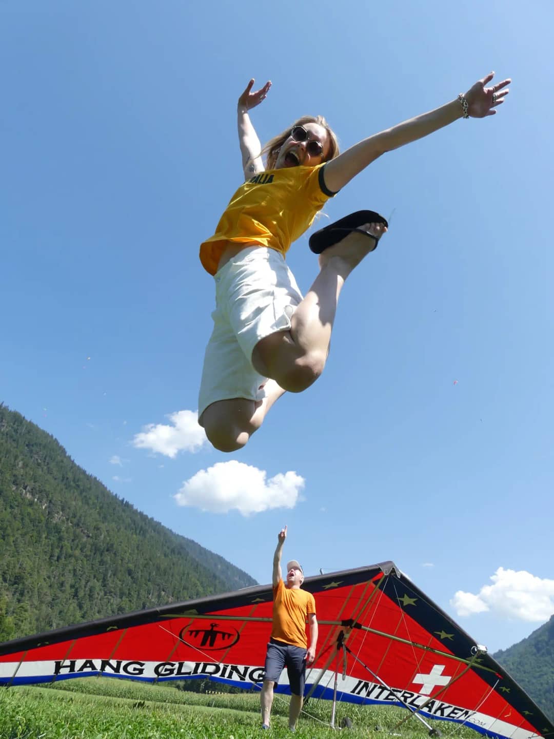 Une femme saute de joie devant un deltaplane rouge Hang Gliding Interlaken posé dans l'herbe, pendant qu'un homme en t-shirt orange la montre du doigt, devant une montagne boisée