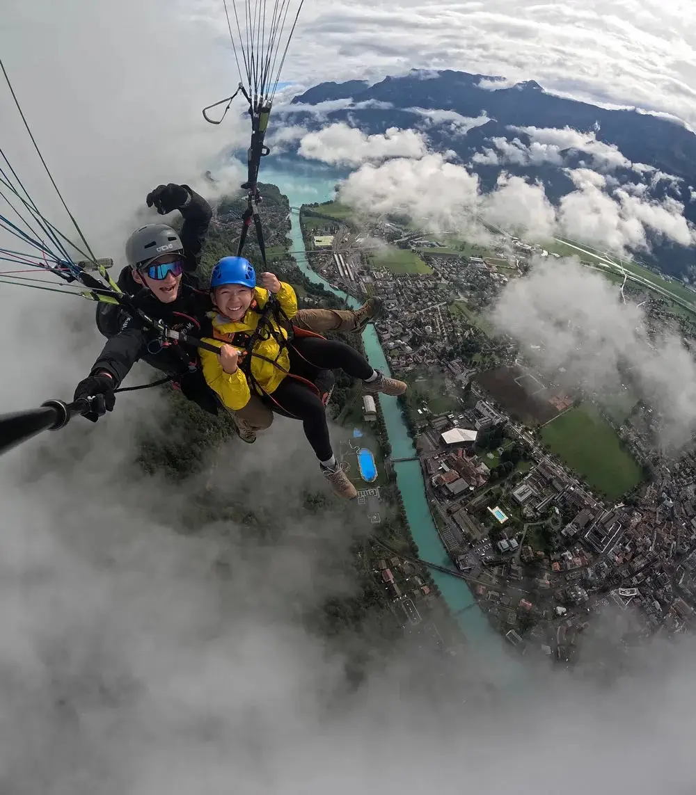 Selfie en parapente biplace au-dessus des nuages : le pilote et une passagère souriante en veste jaune survolent Interlaken, avec l'Aar turquoise qui traverse la ville en contrebas