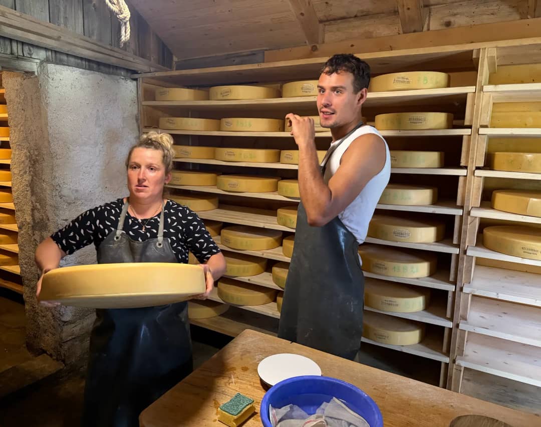 Two alpine cheesemakers in a Justistal cellar lined with ripening wheels of Alpkäse, one holding up a wheel