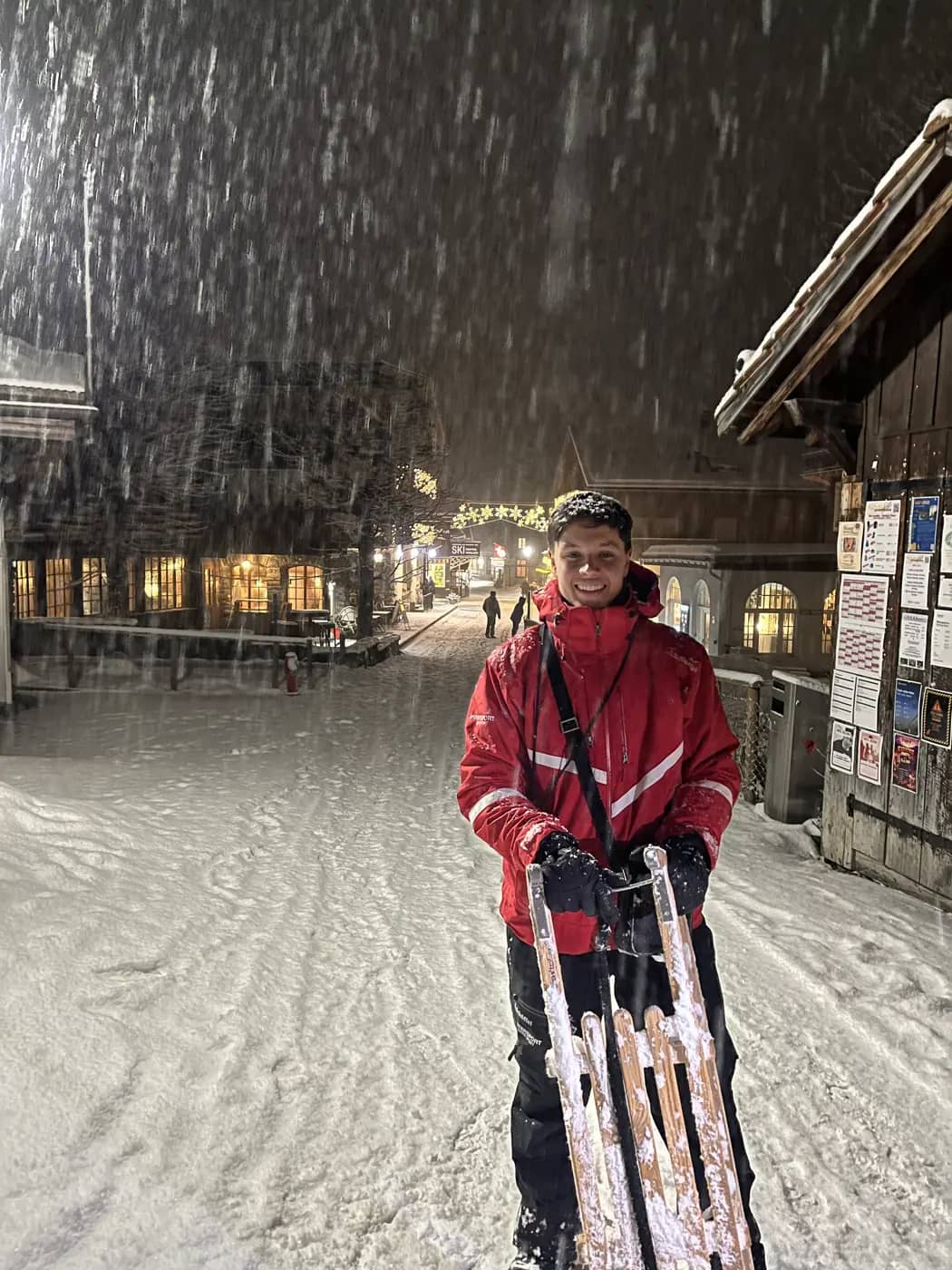 Man holding two wooden sledges in heavy snowfall on the lit main street of Mürren on a January evening