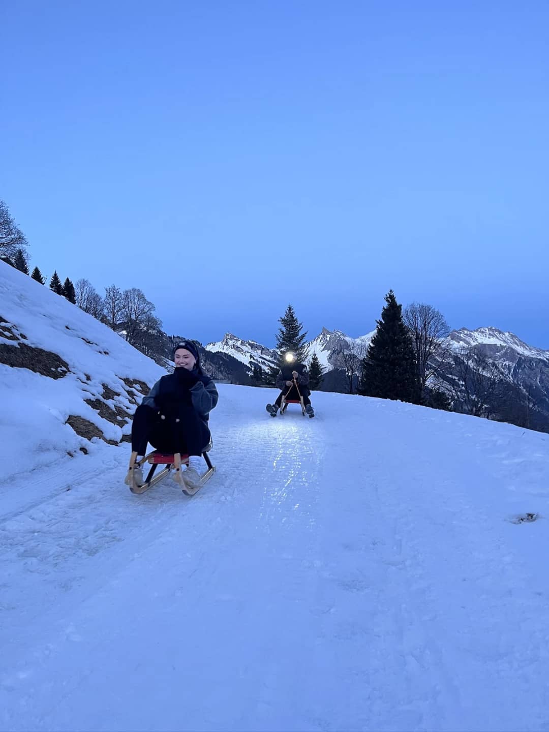 Sledders on wooden toboggans ride down the snowy forest road from Sulwald towards Isenfluh on a clear day