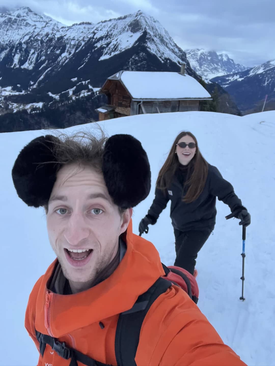 Pierre and a snowshoer with poles climb past a snowed-in wooden chalet on the Sulwald snowshoe trail in winter