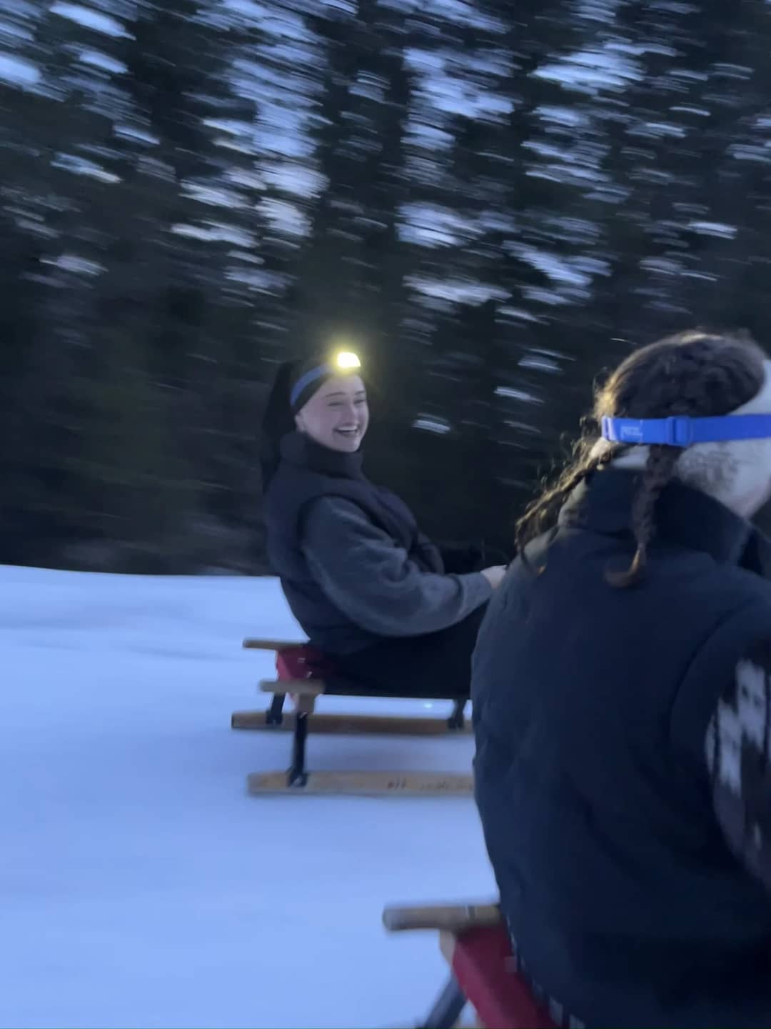 Two sledders with headlamps sit on wooden sleds at dusk on a night sledding Interlaken tour at Isenfluh