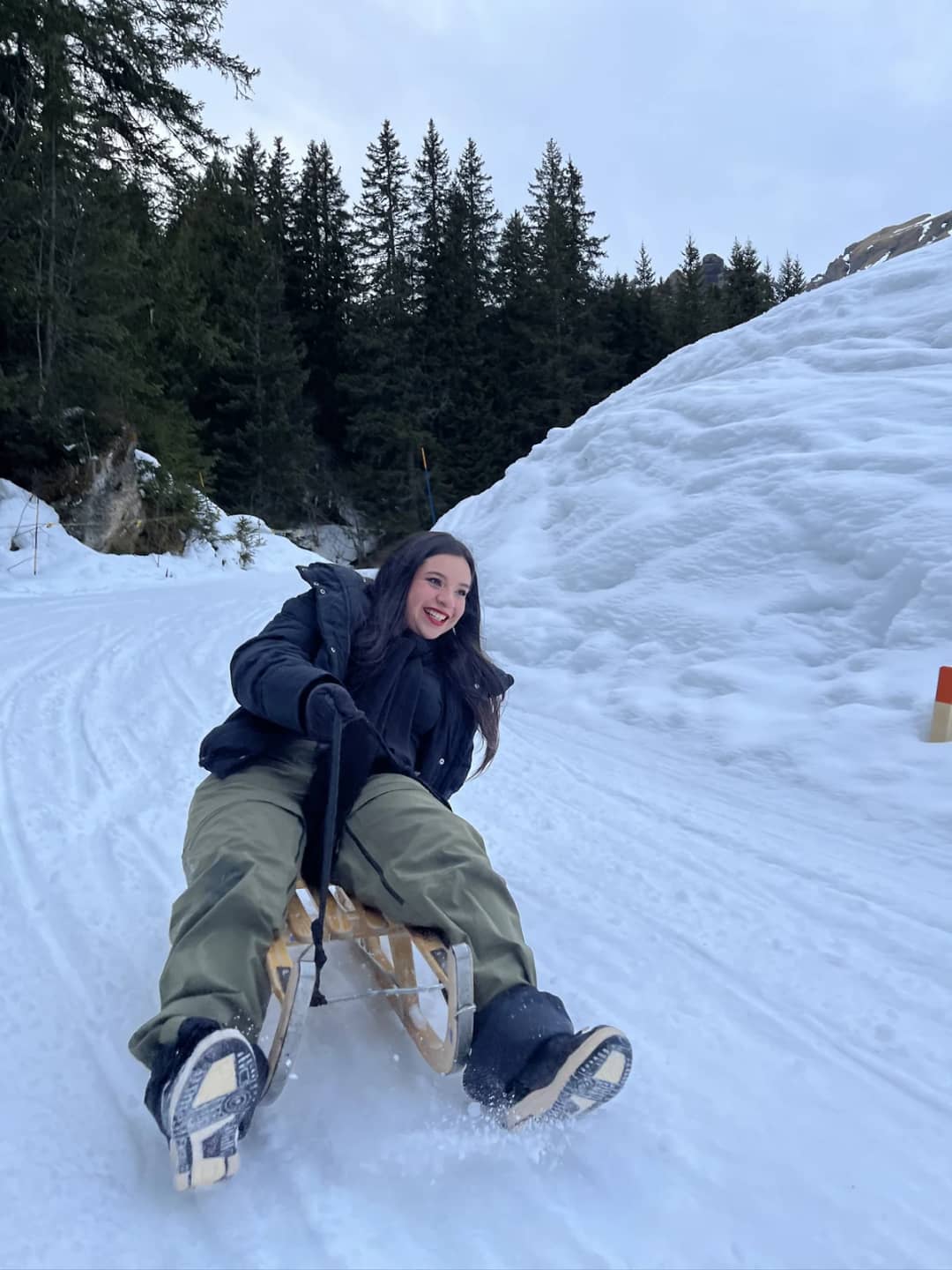 A woman rides a wooden sled down a groomed track between snow banks above Mürren, feet ready to brake