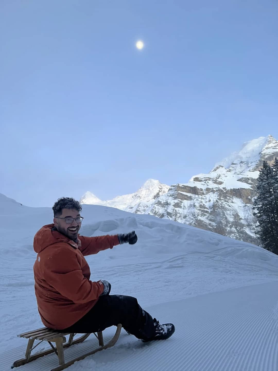 A sledder in an orange jacket sits on a wooden sled at dusk above Mürren, the moon up before night sledding