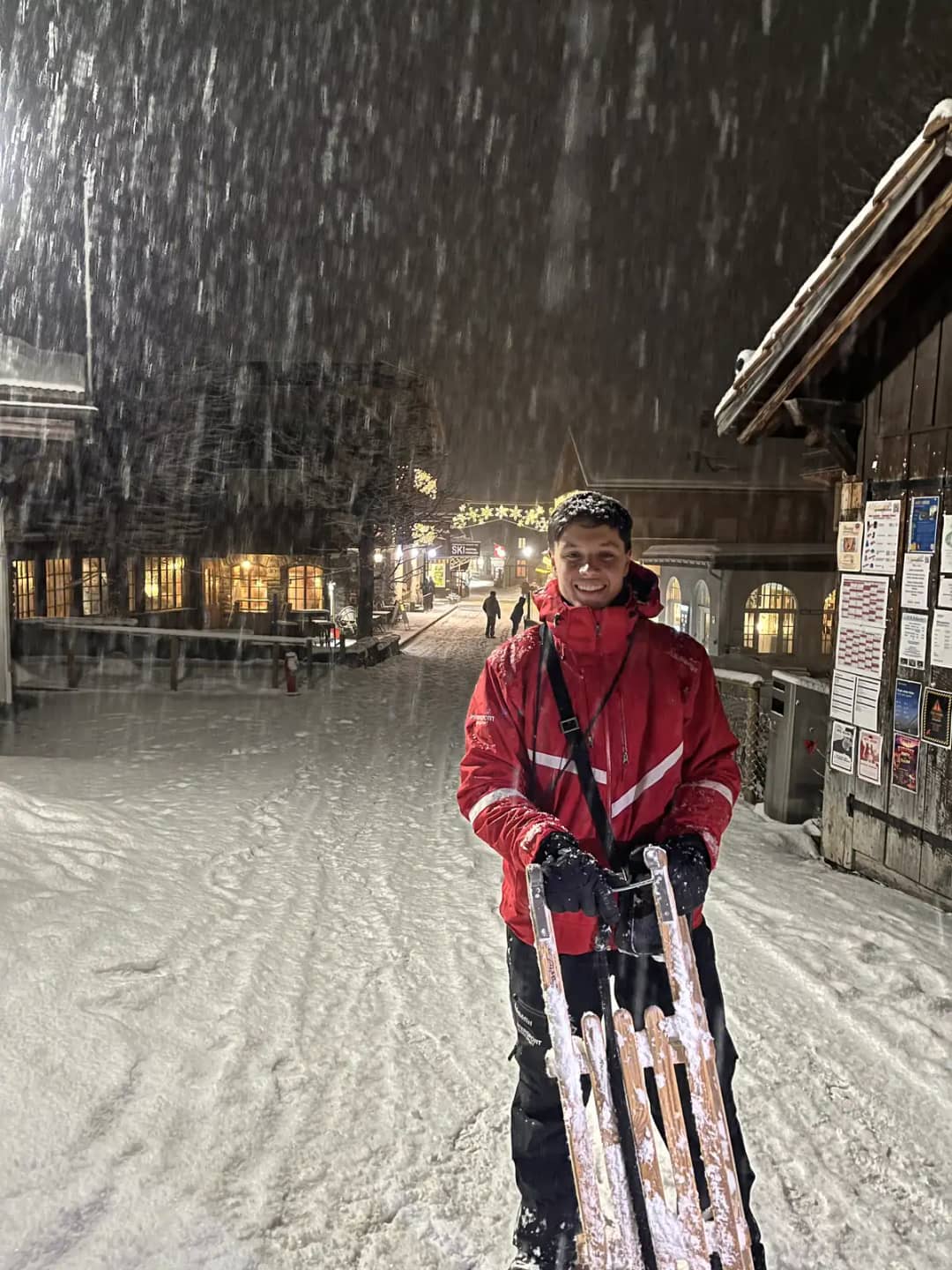 A man in a red jacket holds a wooden sled in the street under heavy snowfall, sledding in Mürren at night