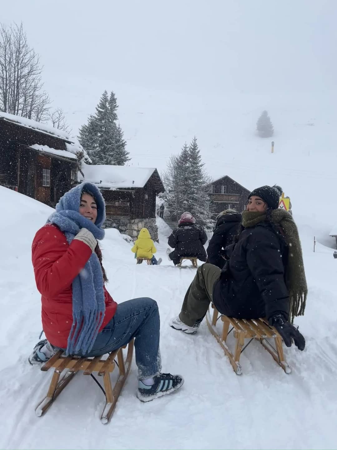 Two sledders sit on wooden sleds between the chalets of Mürren in falling snow on a guided sledding tour