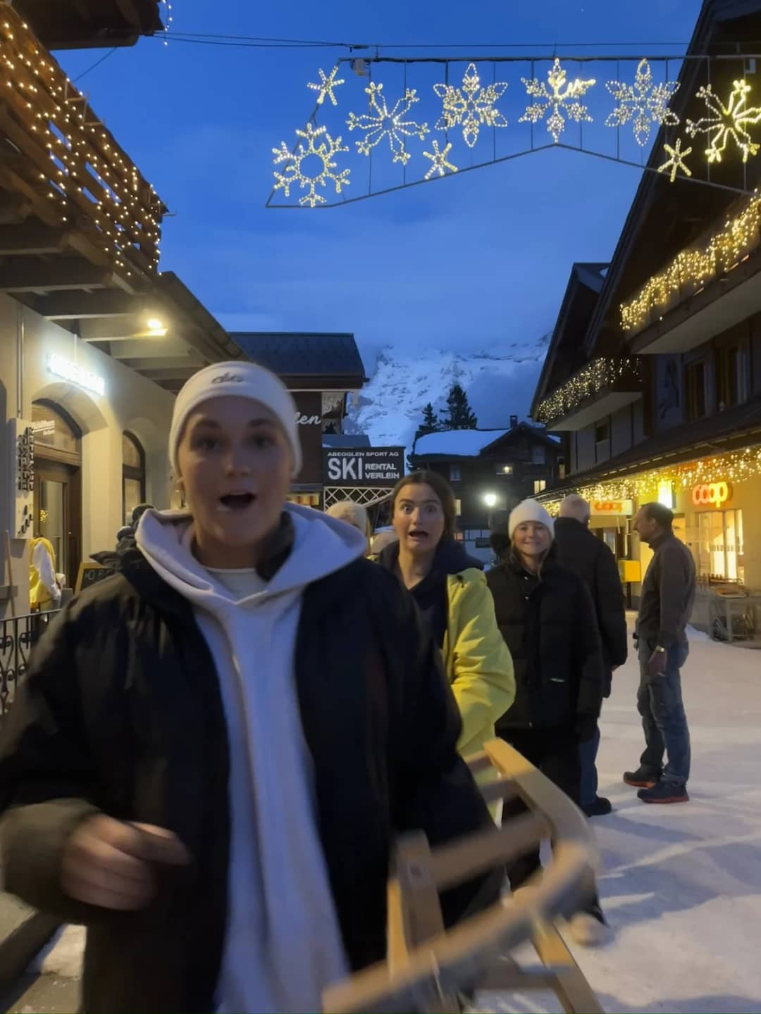 A guest carries a wooden sled through the lit main street at dusk after an evening of sledding in Mürren
