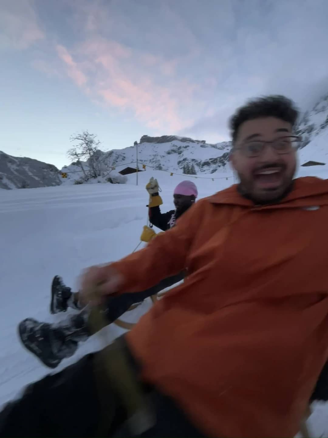 A guest laughs as he speeds down the Mürren sled run on a wooden sled under a pink evening sky