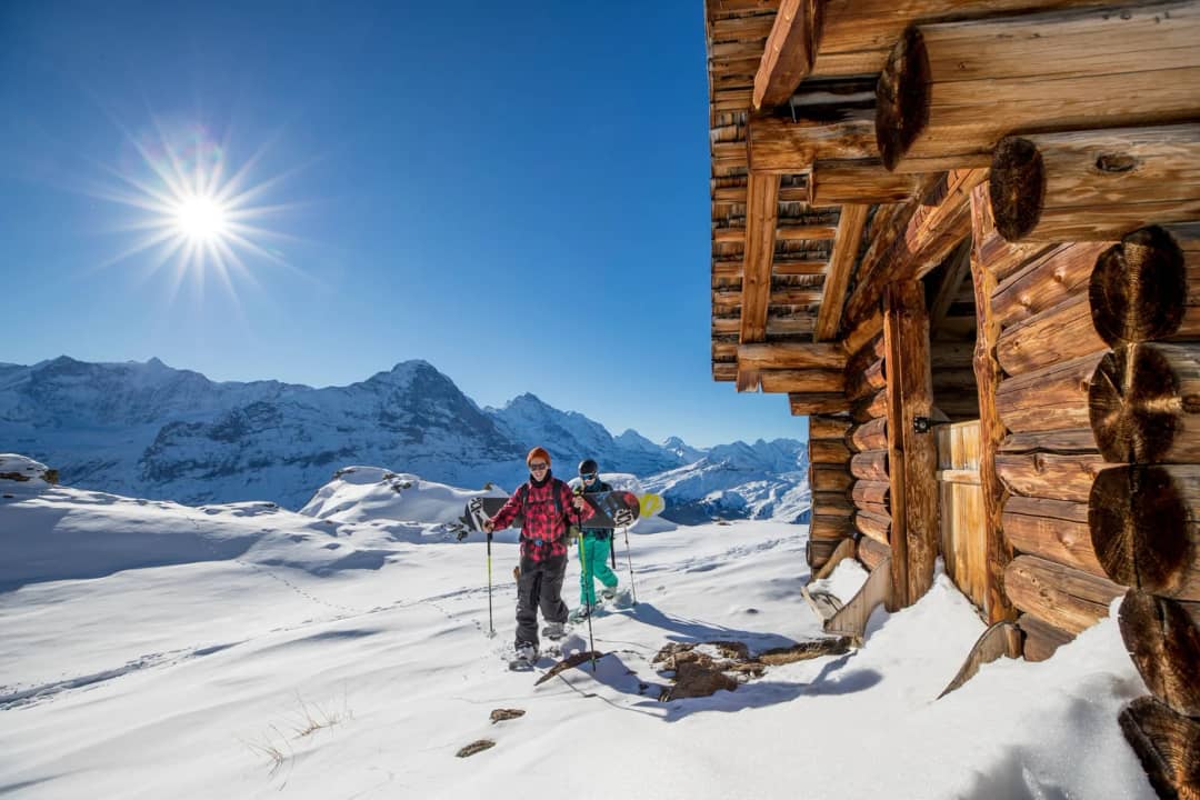 Dos personas con raquetas, una con un snowboard, pasan junto a una cabaña de troncos al sol sobre Grindelwald