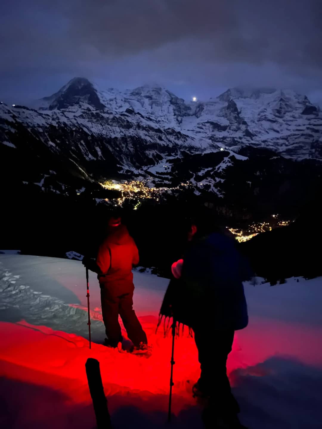 Dos personas con raquetas bajo la luz roja de frontales sobre un valle iluminado y cumbres, raquetas de noche