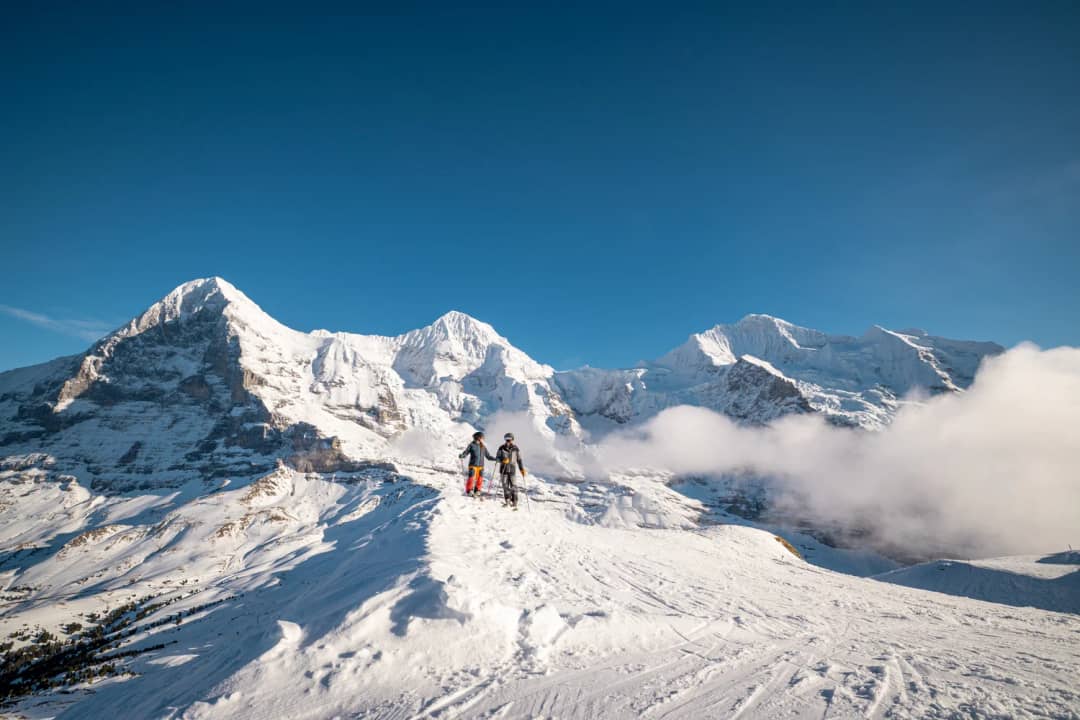 Two skiers stand on a snowy ridge facing the Eiger, Mönch and Jungfrau, with clouds drifting below the peaks.