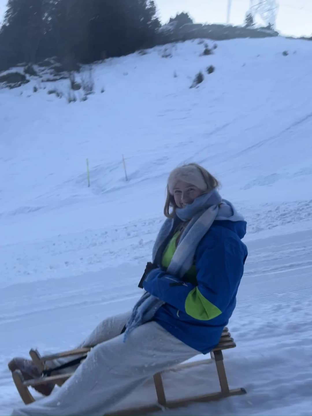 A guest in a ski jacket, a scarf and light snow pants sits on a wooden sled above Mürren in February