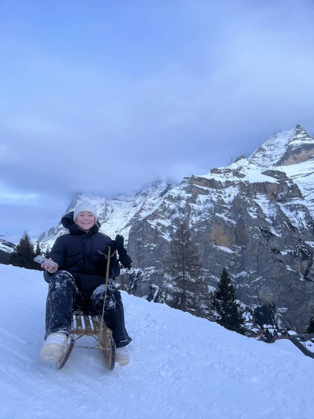 A sledder in high winter boots holds the rope of a wooden sled below the rock walls of Mürren in February