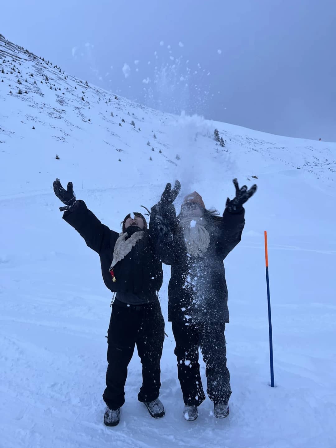 Two guests in black ski clothes, jackets and snow pants, throw powder in the air above Mürren in February