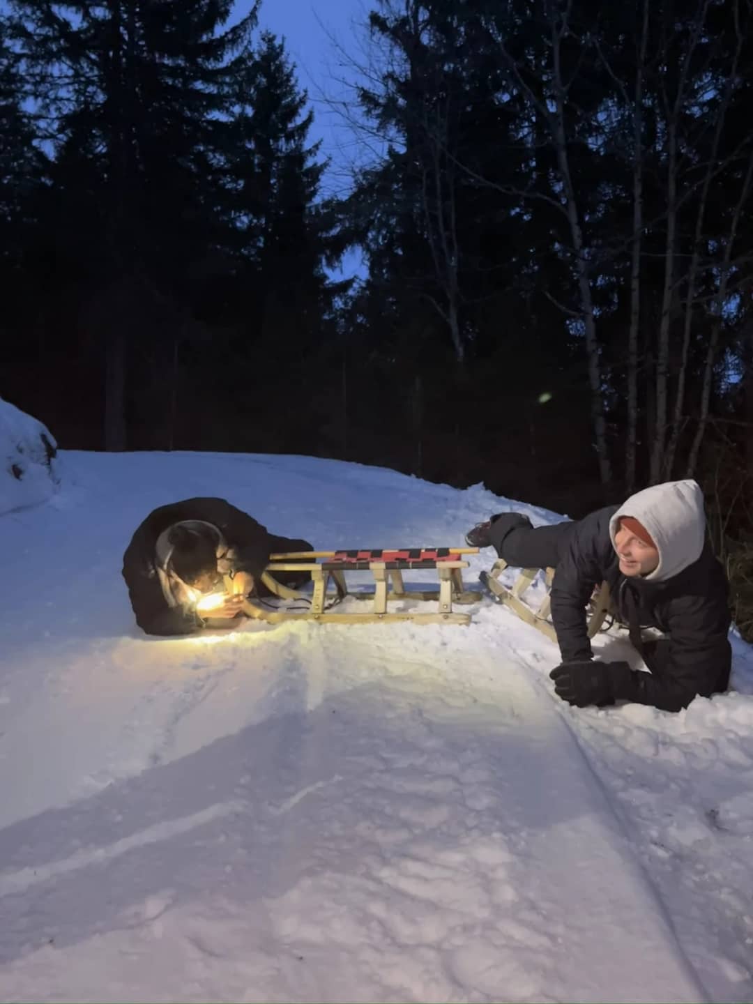 A sledder crouches by two wooden sleds lit by a headlamp on the forest road to Isenfluh on a January evening