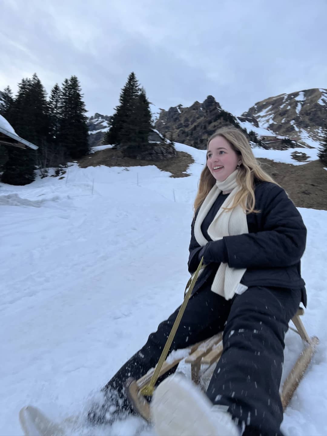 A sledder in a long padded coat and wool scarf kicks up snow spray while braking a wooden sled above Mürren