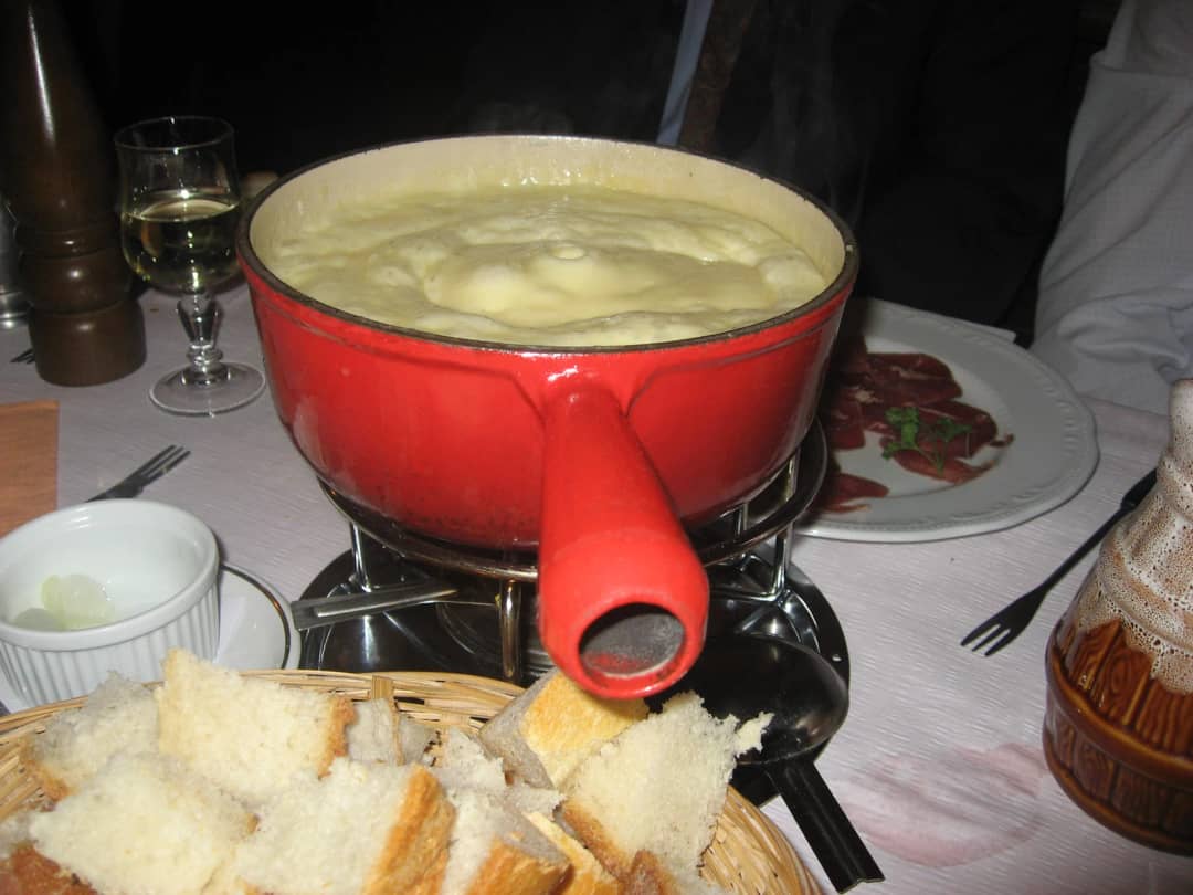Une fondue au fromage dans un caquelon rouge, avec une corbeille de pain en dés et un verre de vin blanc